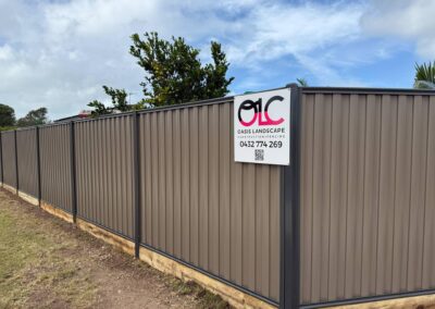 Brown corrugated metal construction fence with an Oasis Landscape sign, phone number and QR code; a fruiting tree is visible behind the fence.