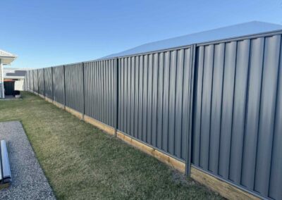 Long gray metal fence running along a grassy backyard under a clear blue sky, with wooden base supports visible at the bottom edge.