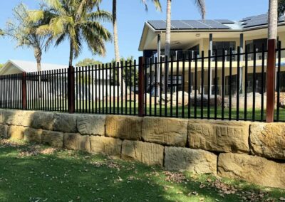 Modern house with solar panels on roof, palm trees, and a black metal fence atop a stone retaining wall.