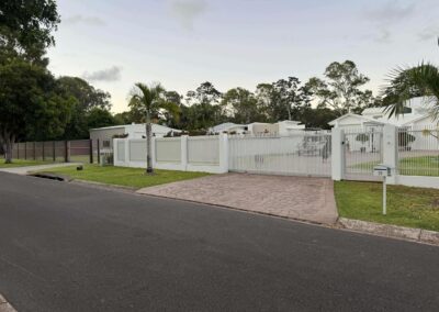 White gated driveway leading to a modern single-story home, with a white fence, palm trees, and a numbered mailbox reading 19.