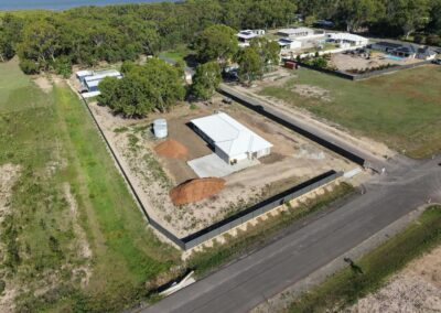 Aerial view of a white, single-story building inside a fenced lot with mulch piles, a water tank, and a gravel drive, surrounded by trees and nearby structures.