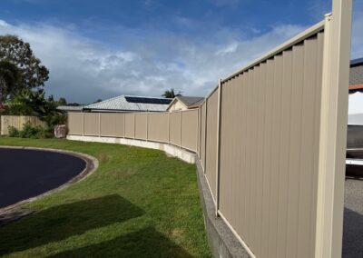 Tall beige corrugated metal fence runs along a curved sidewalk, with trimmed green grass and trees to the left under a blue, partly cloudy sky.