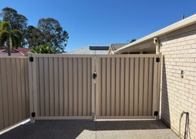 Open driveway gated entrance with beige corrugated metal fence, cream brick wall, and houses with tiled roofs under a blue sky.