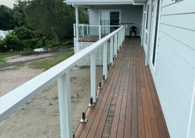Long wooden balcony along a light blue house with white glass-panel railing and a chair at the far end beyond a sliding door.