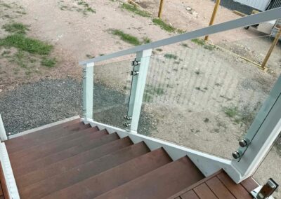 View down a wooden deck staircase with a glass railing and metal posts, opening to a gravel yard outside.