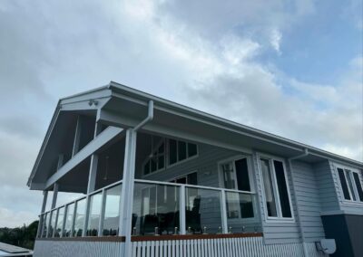 Modern two-story house with a glass-railed balcony, light blue siding, and a cloudy sky.