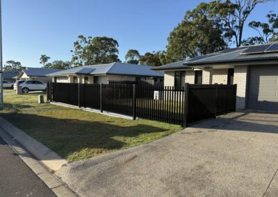 Suburban street scene with single-story brick houses, metal roofs, a tall black fence, and a white car parked along the curb under a clear blue sky