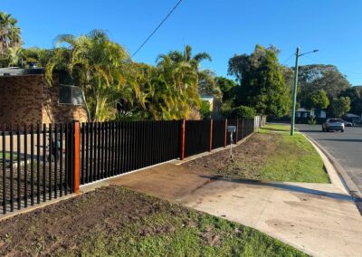 Residential street scene with a tall black fence, palm trees, brick house, and a blue, clear sky; a parked car visible in the distance.