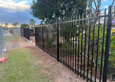 Black metal security fence runs along a dirt path, with temporary construction fencing and stacked materials visible to the left under a cloudy sky.