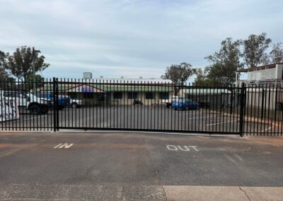 Parking lot behind a tall black metal fence with several cars and a low building in the background under a cloudy sky.