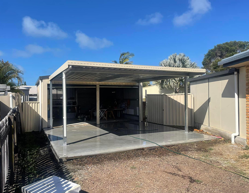 Open-sided metal carport with a concrete patio in a backyard; tools and clutter visible inside, palm trees in the background.