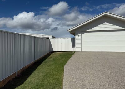White metal fences run along either side of a driveway toward a white garage under a blue, partly cloudy sky.