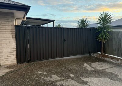 Black metal gate across a driveway with a beige brick wall on the left and tall palm trees in the background under a cloudy sky.