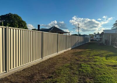 Long beige corrugated metal fence running along a dirt path in a suburban yard, with houses and a blue sky in the background.