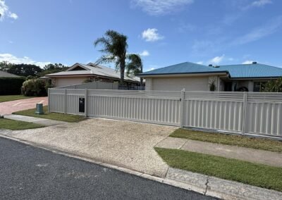 Residential street scene with modern single-story houses, a tall palm tree, and a tall gray metal fence under a clear blue sky with a few clouds.