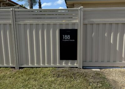 Gray metal fence with a black plaque reading '188 Kingfisher Parade'; palm trees and a teal-roofed house behind.
