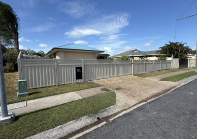 Suburban street with a tall grey metal privacy fence, beige house with teal roof, and a clear blue sky.