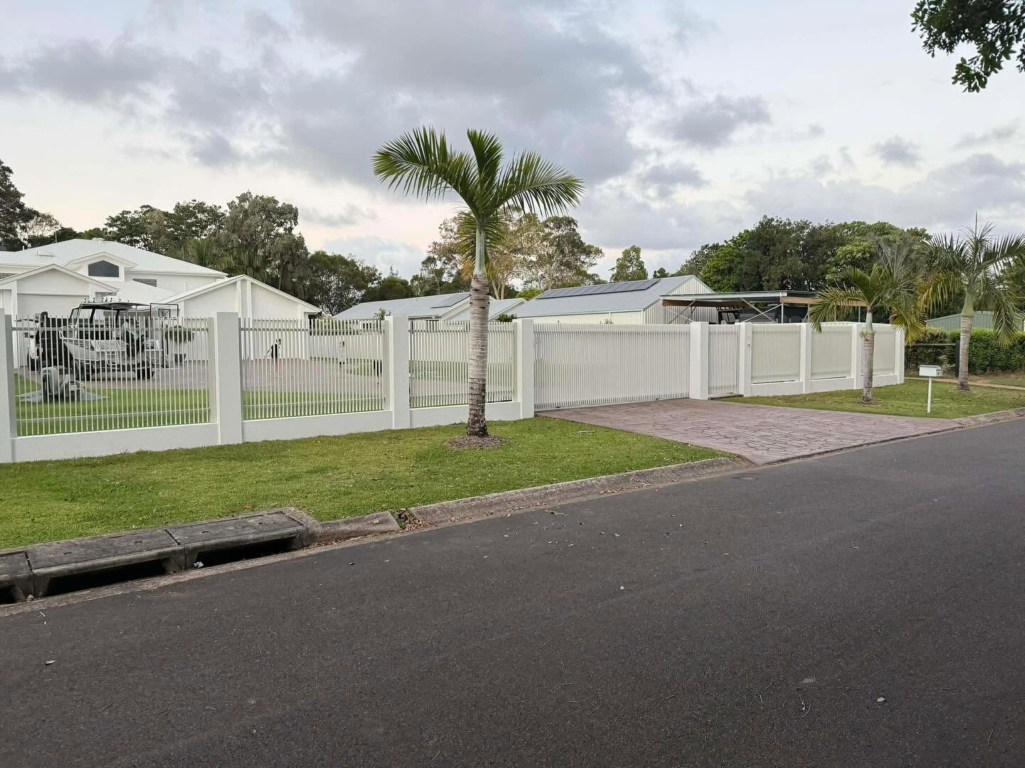 Suburban street scene with white fences, trimmed lawns, and palm trees under a cloudy sky.
