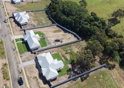 Aerial view of a residential development with white metal roofs, fenced yards, and several cleared dirt lots along a street near a wooded, green area.