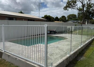 Backyard pool area enclosed by a glass-panel fence on a raised concrete patio, with a cloudy sky and trees in the background.