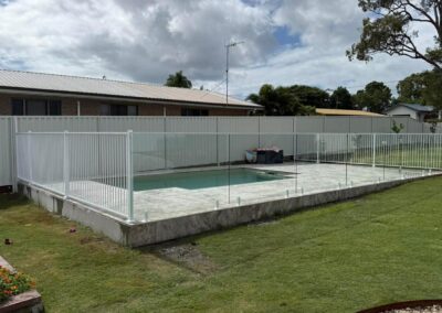 Backyard with a rectangular pool surrounded by a white metal and glass fence on a raised concrete patio.
