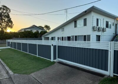 White and blue privacy fence along a lawn with a two-story house and sunset in the background, air conditioning units on the side, and a gate visible at the corner.