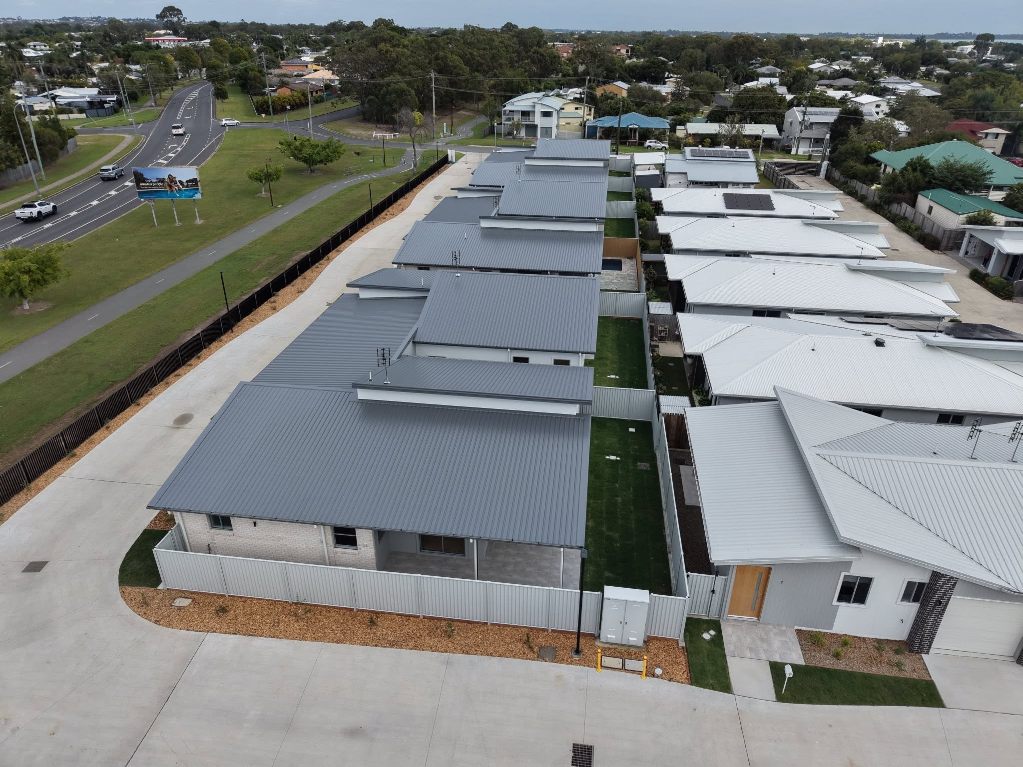 Aerial view of a row of grey-roofed townhouses with driveways, fencing, and a nearby park and road.