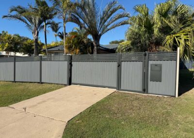 Tall gray privacy fence with a metal gate, palm trees behind it and a clear blue sky above, a concrete sidewalk in the foreground.