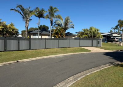 Residential street scene with a tall gray privacy fence, palm trees, and single-story houses under a clear blue sky.