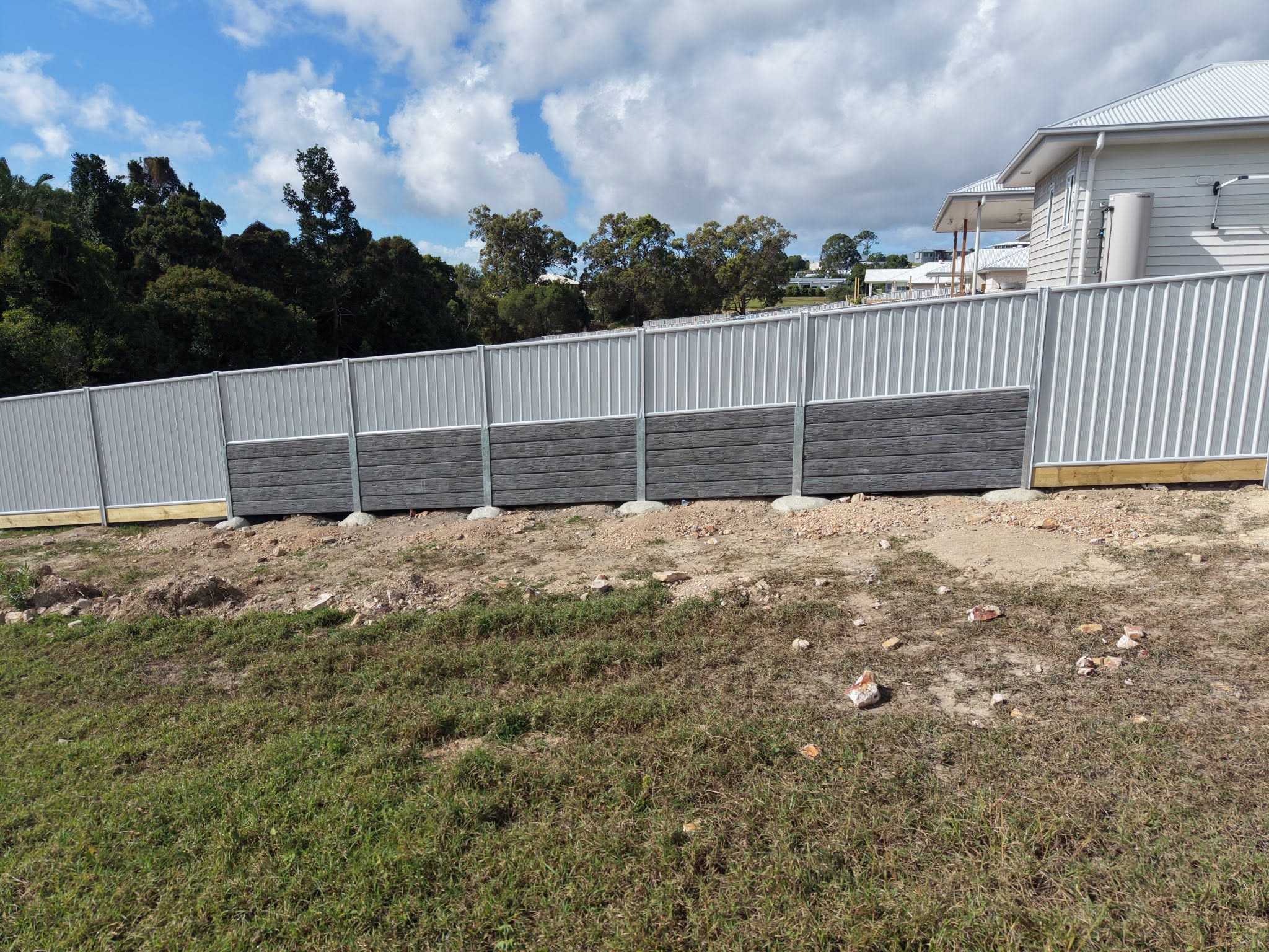 Grey corrugated metal fence lines a dirt lot with trees in the background and a white-roofed house to the right.
