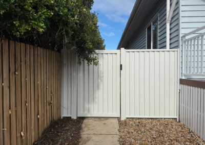 Narrow alley between a wooden fence on the left and a white gate beside a gray house, with gravel and a clear blue sky.