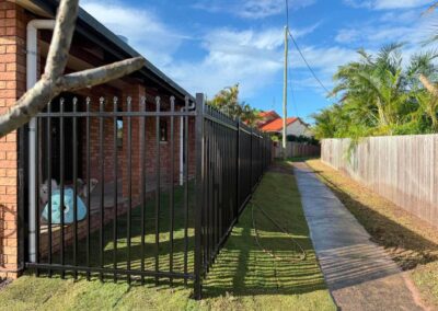 Brick house with a black metal fence, a small white dog near a blue hose in the shaded patio, under a bright blue sky with palm trees nearby.