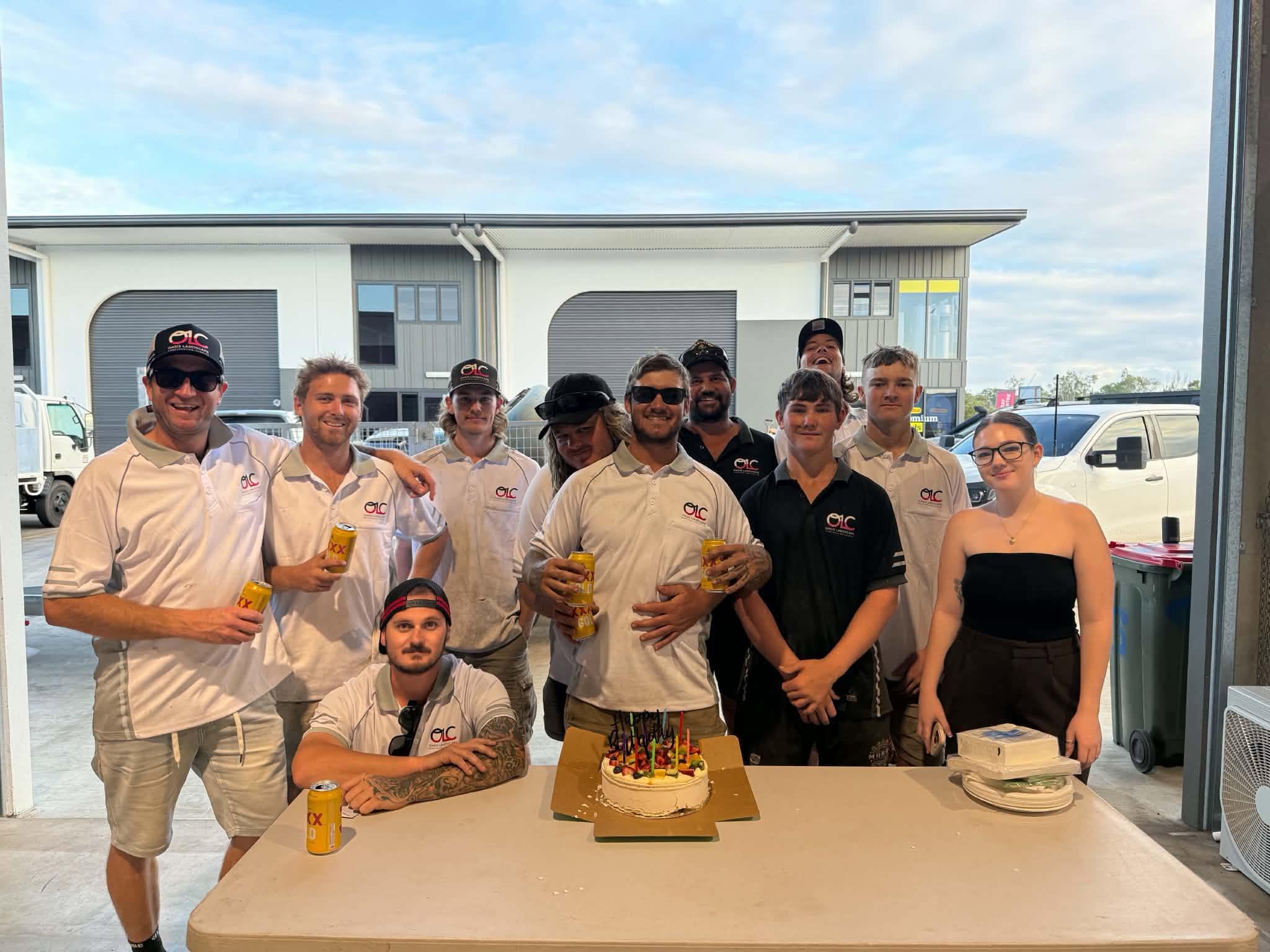 Group of coworkers in matching shirts posing around a birthday cake with candles and canned drinks at a workplace celebration.
