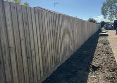 Long wooden privacy fence along a dirt path on a sunny day, with a gravel strip and distant trees/vehicles visible at the end of the fence.