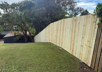 Backyard scene showing a tall pale wooden privacy fence running diagonally across a grassy yard, with trees and a dark shed behind it.