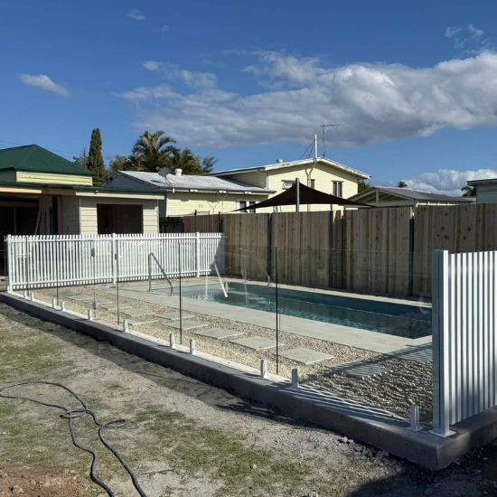Rectangular in-ground pool with metal railing and white fencing in a backyard under a blue sky, with nearby houses beyond the fence.
