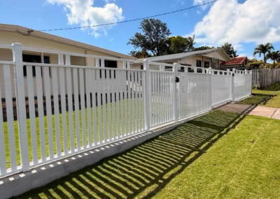 White metal fence with a gate along a sidewalk in front of single-story houses; long shadows on the green lawn.