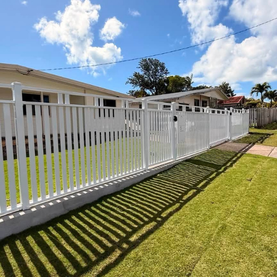 White metal fence with a gate along a sidewalk in front of single-story houses; long shadows on the green lawn.