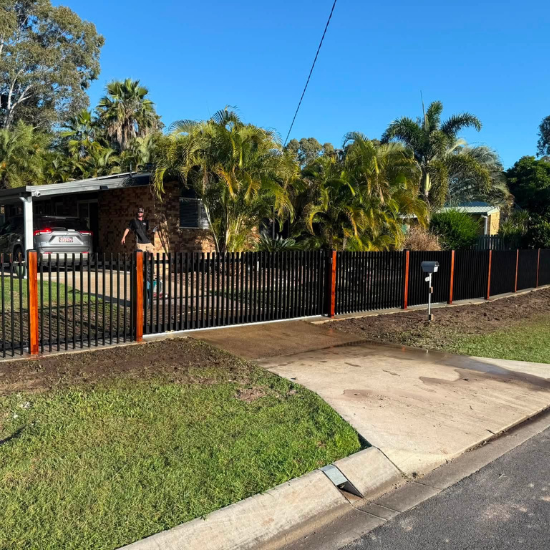Residential front yard with a black metal fence and orange posts, palm trees, a driveway, and a parked car beside a brick house distant to the left.