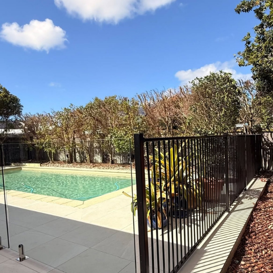 Residential backyard pool with a black metal fence, light-patina deck, and potted plants on a sunny day.