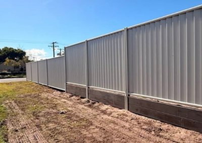 Long row of light gray corrugated metal fence panels along a dirt lot under a clear blue sky.