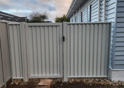 Gray metal privacy gate between fence panels beside a blue house, with a stone stepping path and dirt/rock edging in the yard.