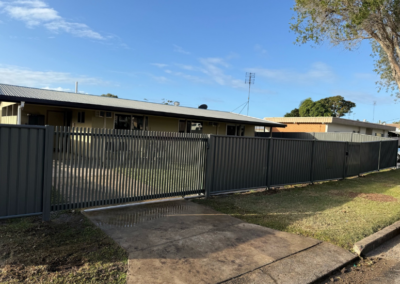 A row of single-story beige houses behind a tall gray metal slat fence with a sliding gate and a concrete driveway in the foreground under a blue sky.