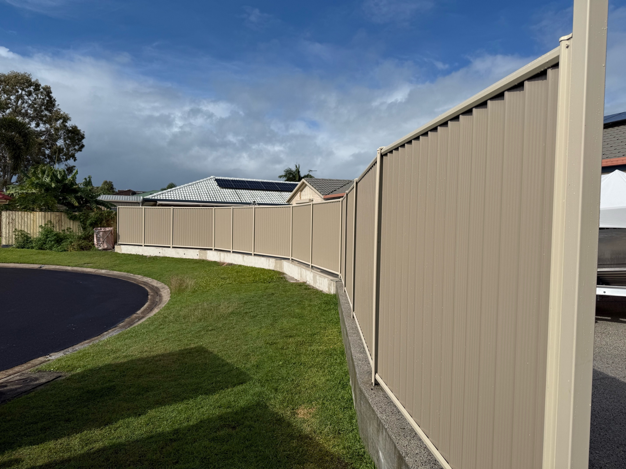 Beige corrugated privacy fence runs along a curved curb in a residential yard, with green grass and neighboring rooftops in view.
