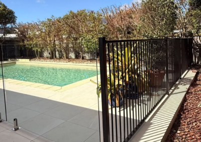 Backyard swimming pool enclosed by a glass panel fence and black metal railing, with greenery along the fence and a clear blue sky above.