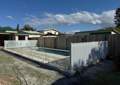 Backyard pool under construction with a glass fence, wooden fence panels, and surrounding houses under a blue sky with clouds.