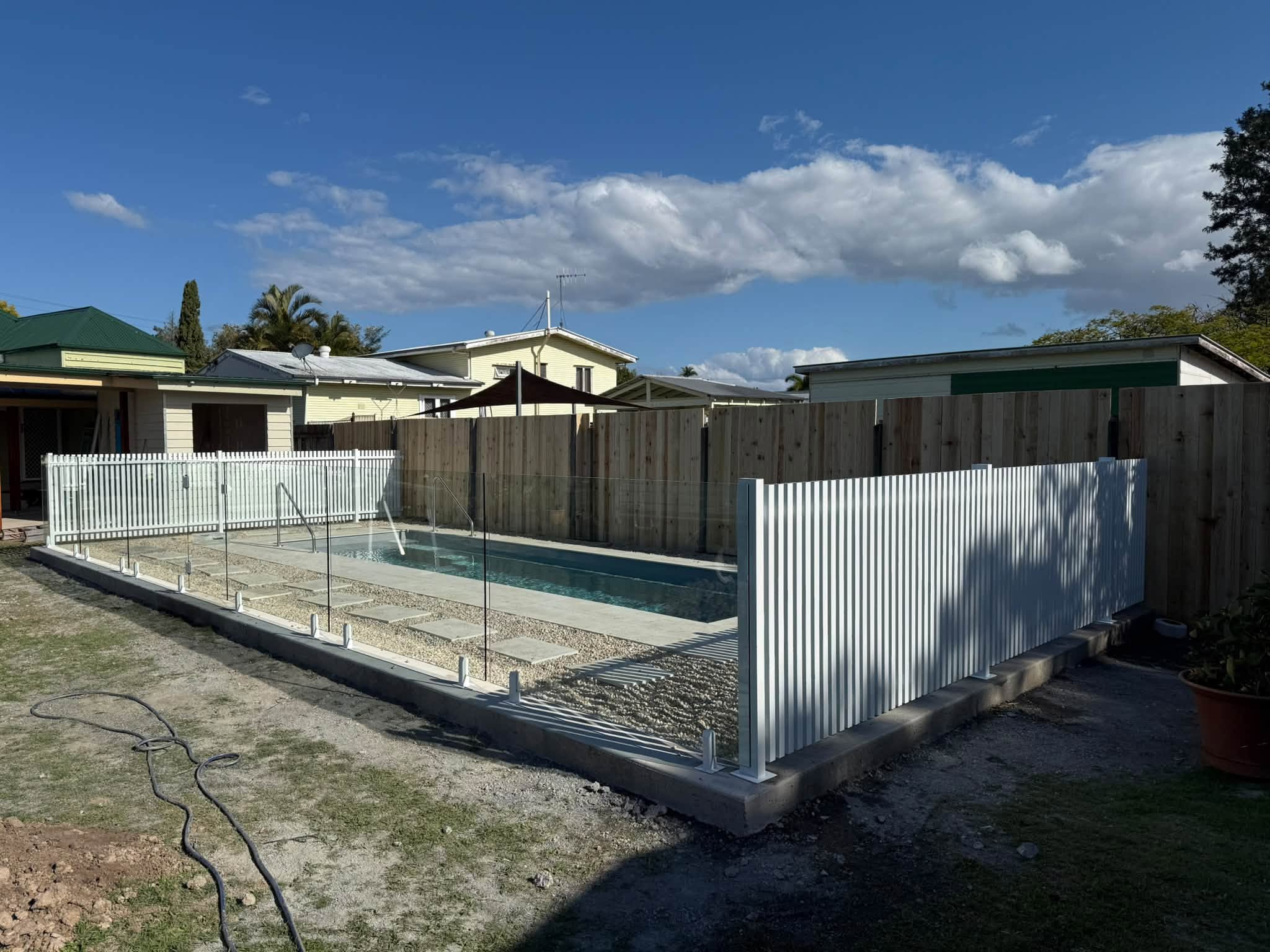Backyard pool under construction with a glass fence, wooden fence panels, and surrounding houses under a blue sky with clouds.