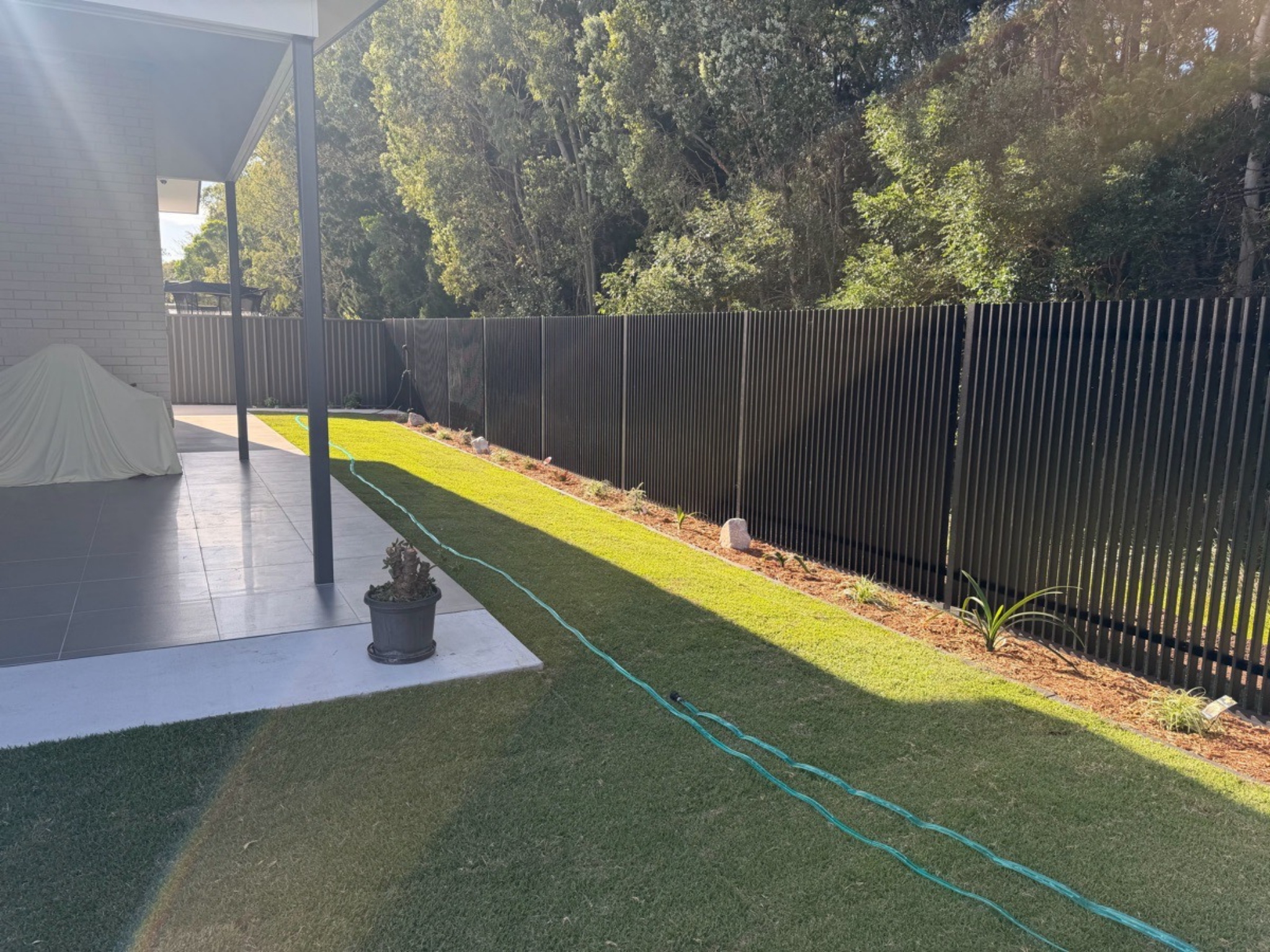 Patio area with a covered structure on the left, a tall black fence, and a manicured lawn with a garden hose laid across.