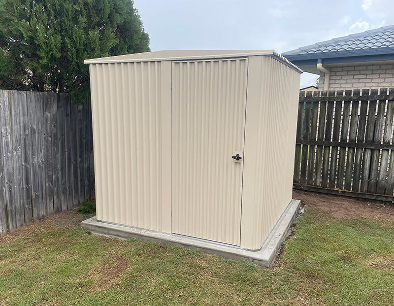 Cream Colorbond garden shed on a concrete slab in a backyard corner, installed by Oasis Landscape Construction and Fencing in Hervey Bay.